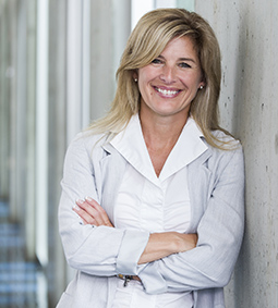 Executive Coach Nicola Johnson, a woman with blonde hair and a white blouse stands with her arms crossed, smiling confidently. She is wearing a light grey blazer and leaning against a wall in a corridor with blurred background.